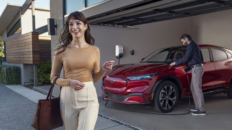 A woman walks away from a Mustang Mach-E as a man plugs it into a home charging station